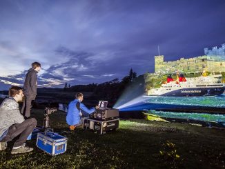 A large projector on a grass verge at the side of a loch surrounding Dunvegan Castle, clouds are moving overhead as the sun sets. Behind the projector a man is crouched using a laptop.The machine is projecting a beam of light across the water which is projecting the image of a Caledonian Macbrayen Ferry on to the side of the castle. Another man is crouched behind a camera and slider aparatus while a third man stands to the left of the projector looking towards the castle.